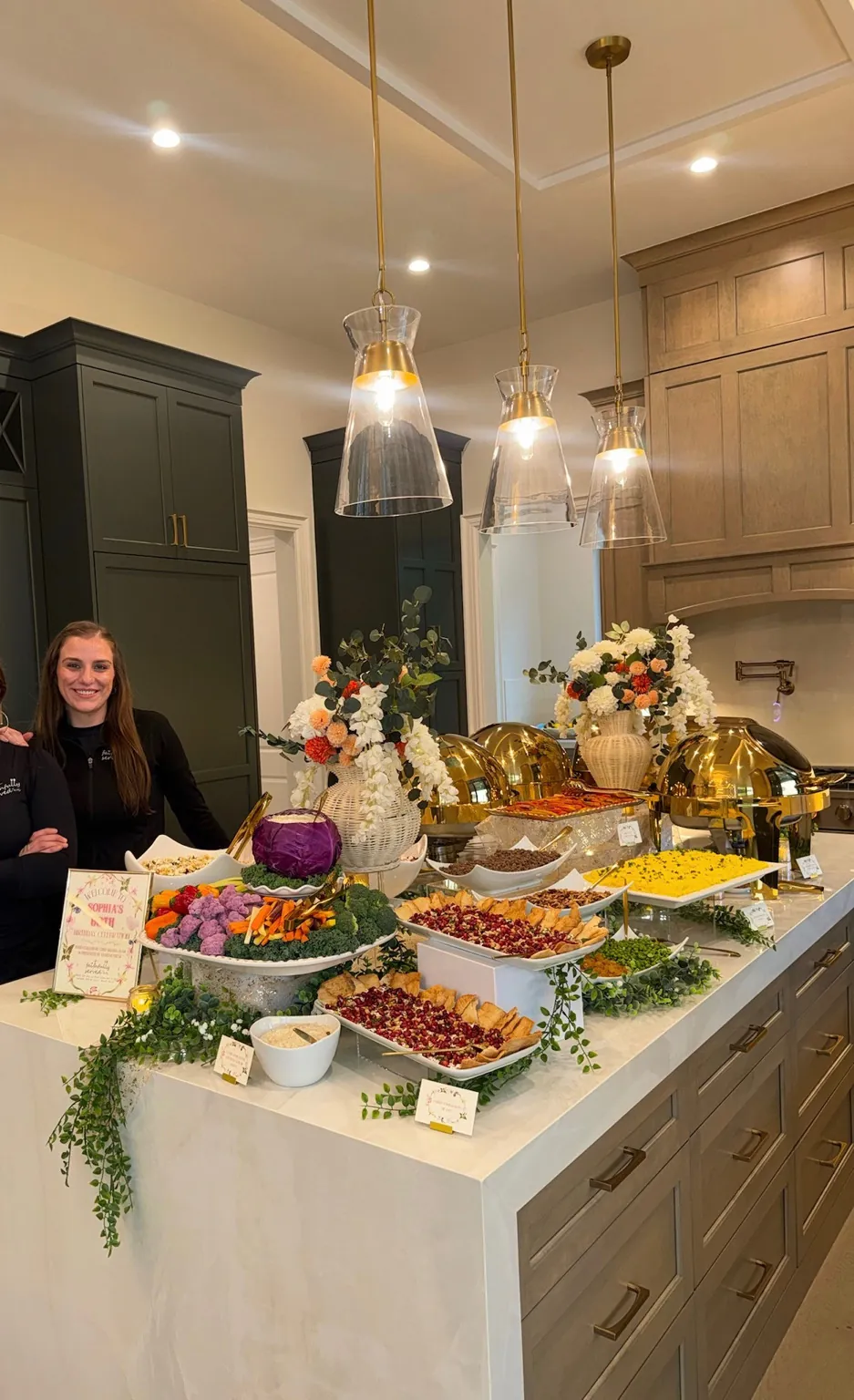 Mariah beside a Gracefully Served presentation — gold chafing dishes, crudité boards and florals styled along a kitchen island